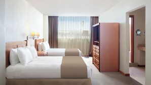 Habitación de hotel con dos camas, un armario de madera, un escritorio y un ventanal grande con cortinas.