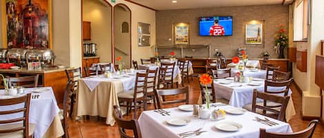 A dining area with multiple tables set for guests, a buffet station, and a television mounted on the wall.
