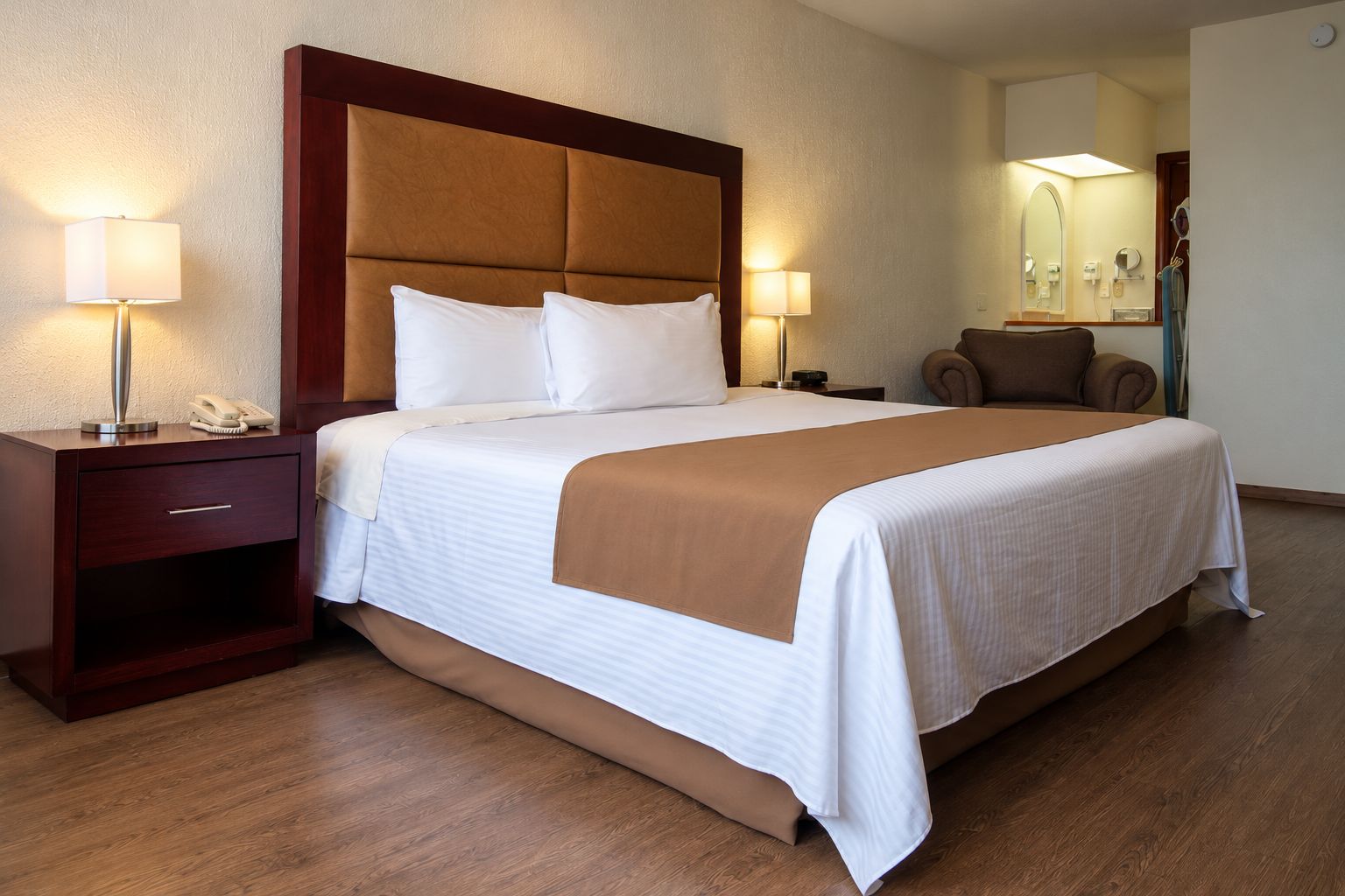 Una habitació d'hotel amb un llit gran, taules de nit, una cadira, un mirall i una làmpada.