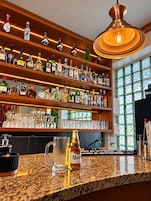 Zona de bar con estantes llenos de botellas de licor, una lámpara colgante de cobre y una encimera de granito con una botella de vino Modena.