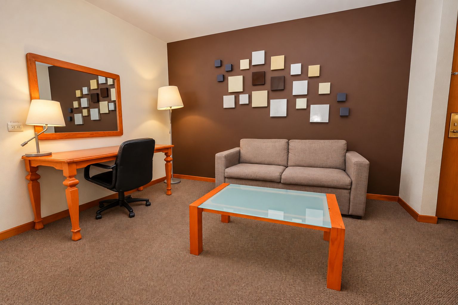 Habitación de hotel con escritorio, dos sillas, un sofá, una mesa de centro de vidrio, un espejo en la pared y una obra de arte en la pared.