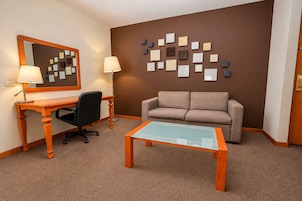 Habitación de hotel con escritorio, dos sillas, un sofá, una mesa de centro de vidrio, un espejo en la pared y una obra de arte en la pared.