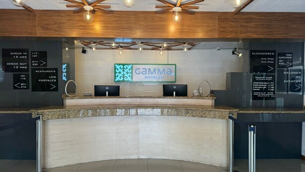 Zona de recepción de hotel con diseño moderno, que incluye un mostrador de recepción, un cartel de Gamma Hotels, indicaciones para el registro de entrada y salida, y un techo con un diseño de estrellas de madera y luces colgantes.