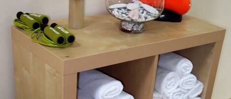 A wooden shelf with neatly rolled white towels, a glass vase with decorative items, and exercise equipment including yoga mats and dumbbells.