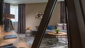 A modern hotel room with a wooden desk, a laptop, a telephone, a small table with a plant, a sofa with cushions, and a decorative wall piece.