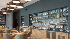 Un caffè moderno con tavoli e sedie in legno, un bancone con cibo e bevande, e scaffali con bottiglie e oggetti decorativi.