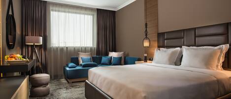 A modern hotel room with a large bed, a seating area with blue sofas, a desk with a fruit bowl, and a large window with curtains.