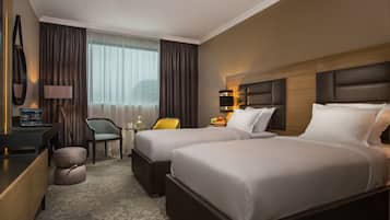 Quarto de hotel com duas camas, uma escrivaninha, uma cadeira, uma mesinha de cabeceira com abajur e uma janela com cortinas.