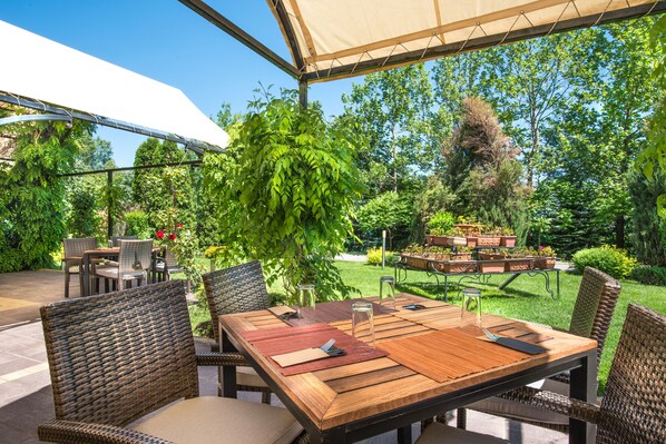 An outdoor patio with a wooden table and wicker chairs, surrounded by lush greenery and a white canopy.