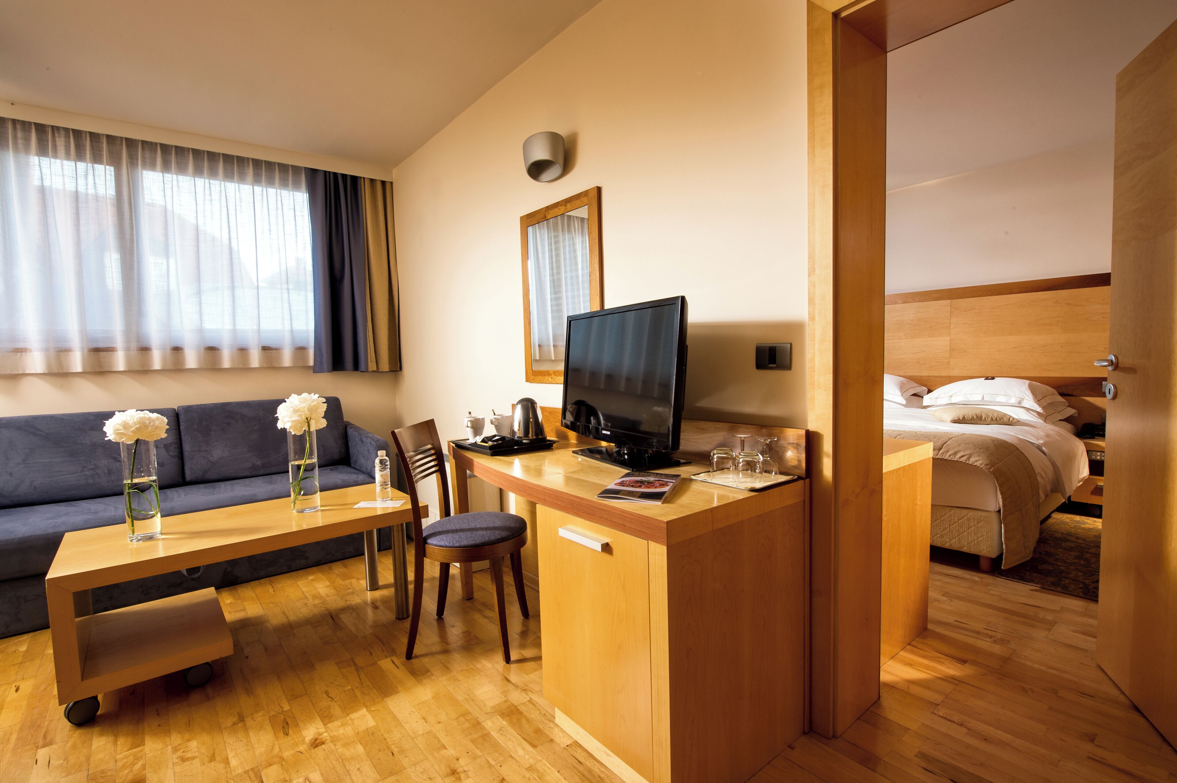A modern hotel room with a wooden desk, a television, a sofa, and a bed.