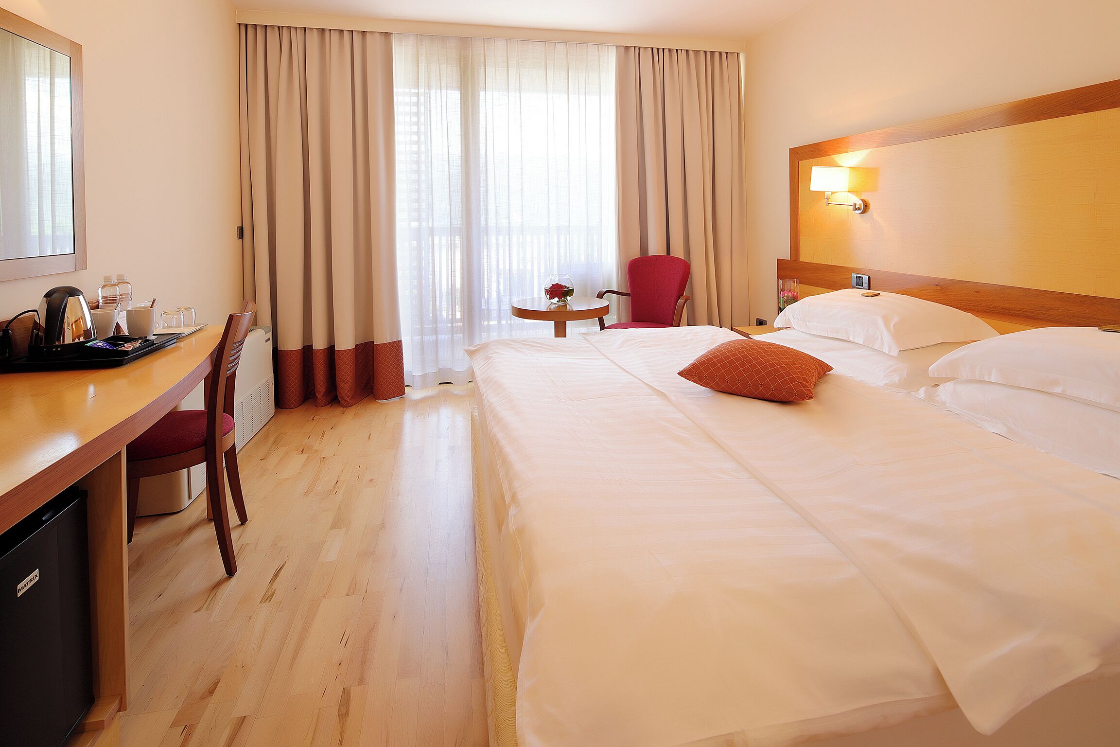 A hotel room with a bed, a desk with a chair, a small table with a vase, and a window with curtains.