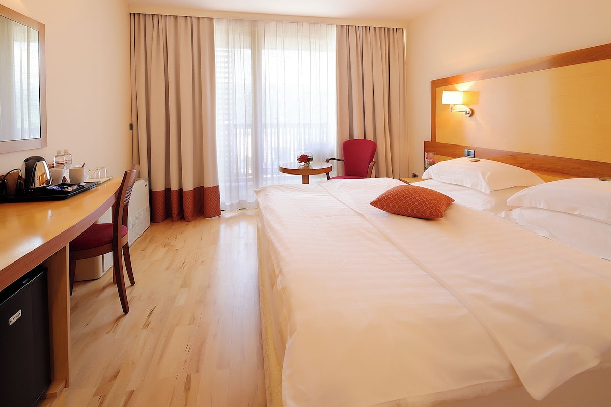 A hotel room with a bed, a desk with a chair, a small table with a vase, and a window with curtains.