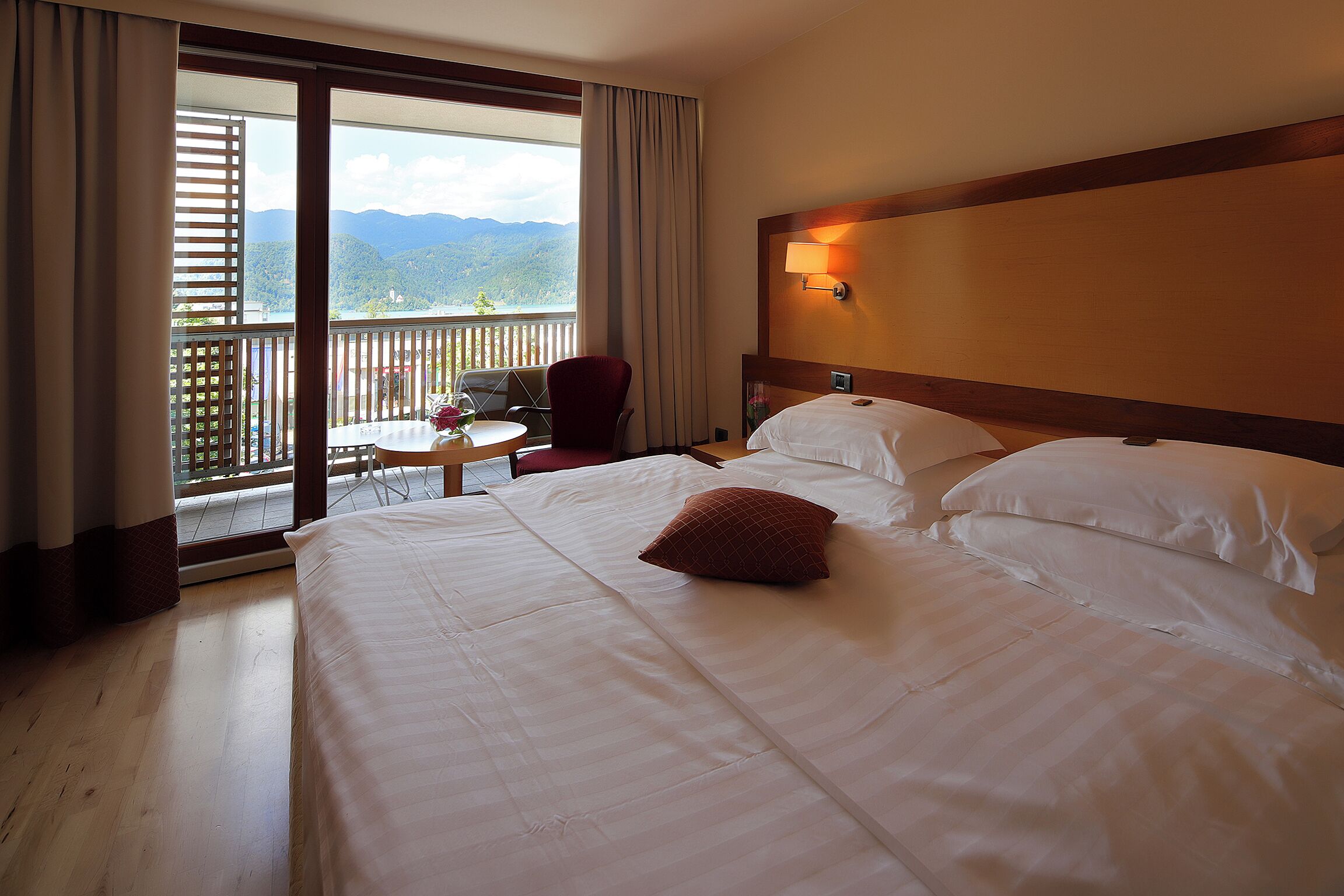 A hotel room with a large bed, a chair, a small table, and a balcony with a view of mountains.