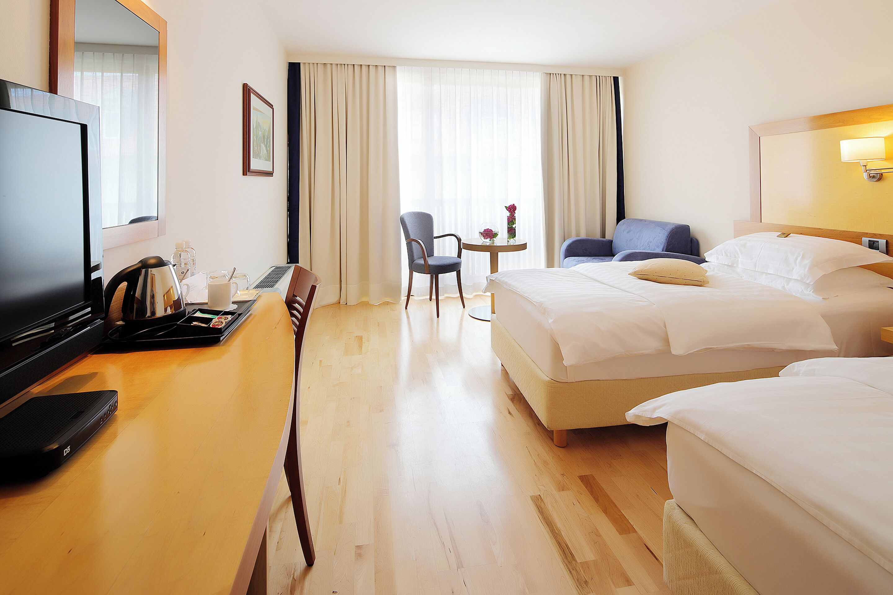 A hotel room with a bed, a desk with a laptop, a chair, a TV, and a window with curtains.