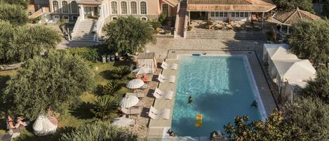 Vista aérea de um resort com piscina, espreguiçadeiras e um edifício grande com telhado de azulejos.