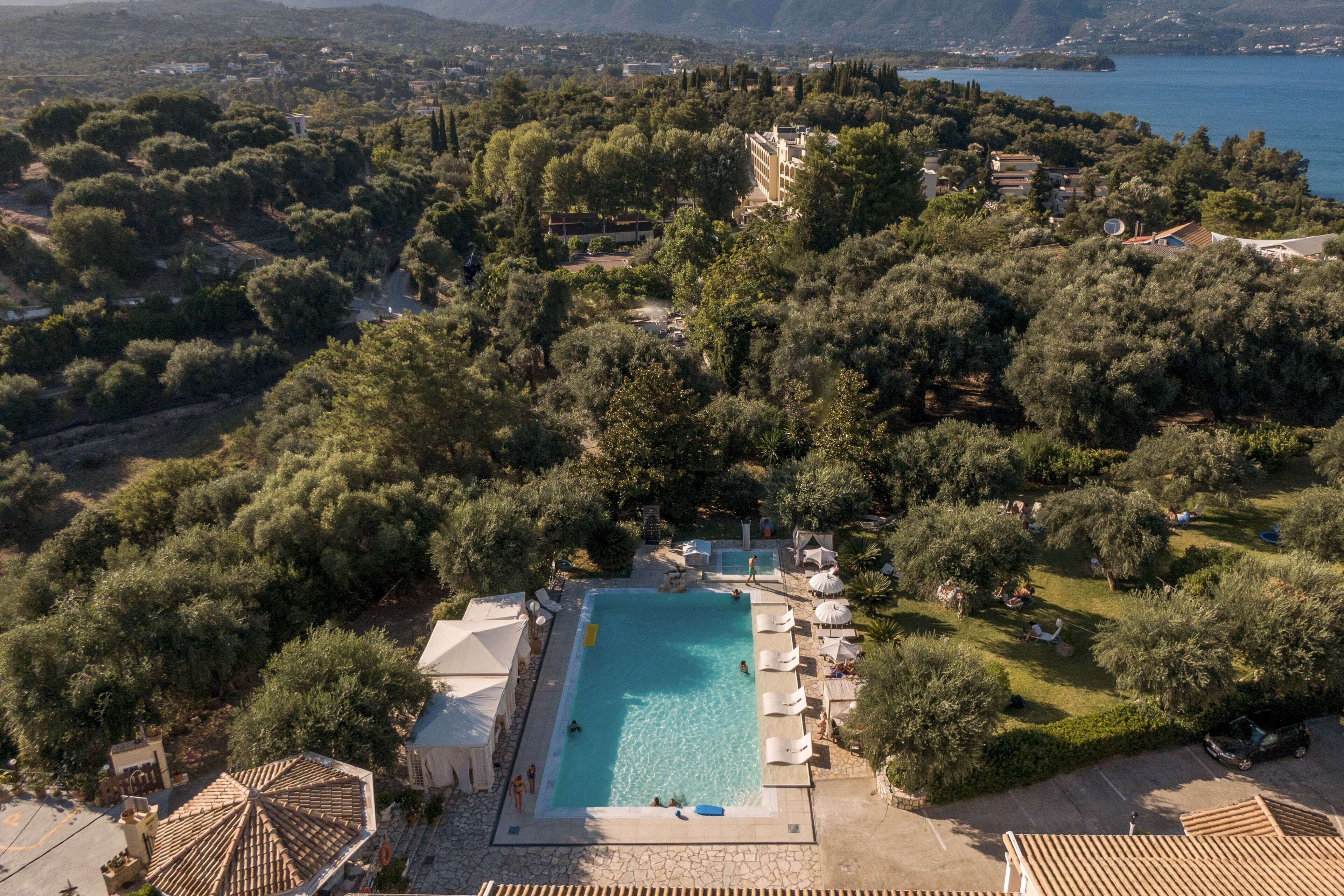 An aerial view of a resort with a swimming pool surrounded by lush greenery and a view of the sea.