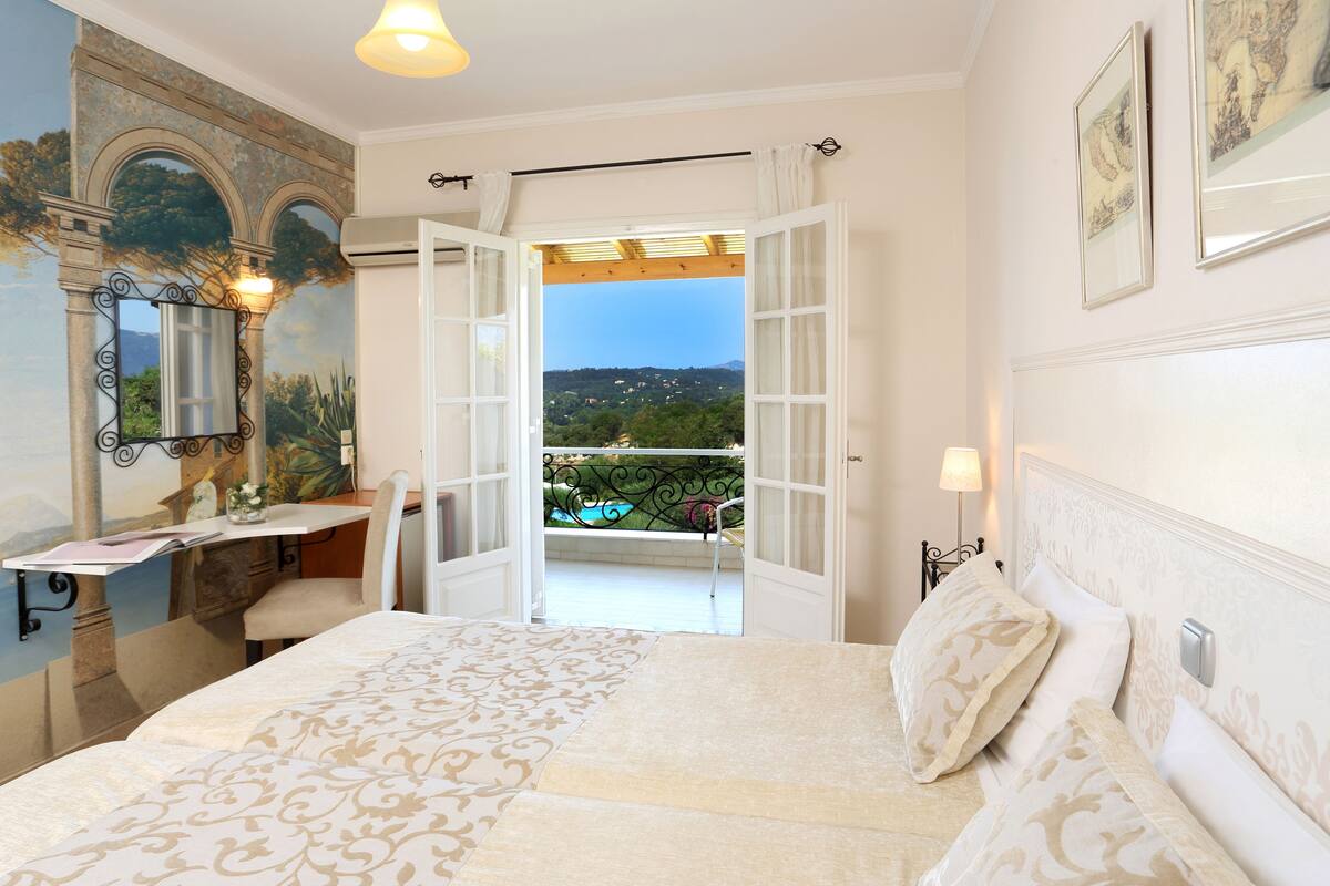 A bedroom with a bed, a desk, a chair, a mirror, and a view of a balcony with a pool.