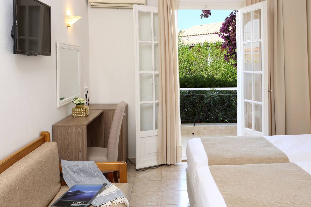 A hotel room with a bed, a desk, and a view of a balcony with plants.