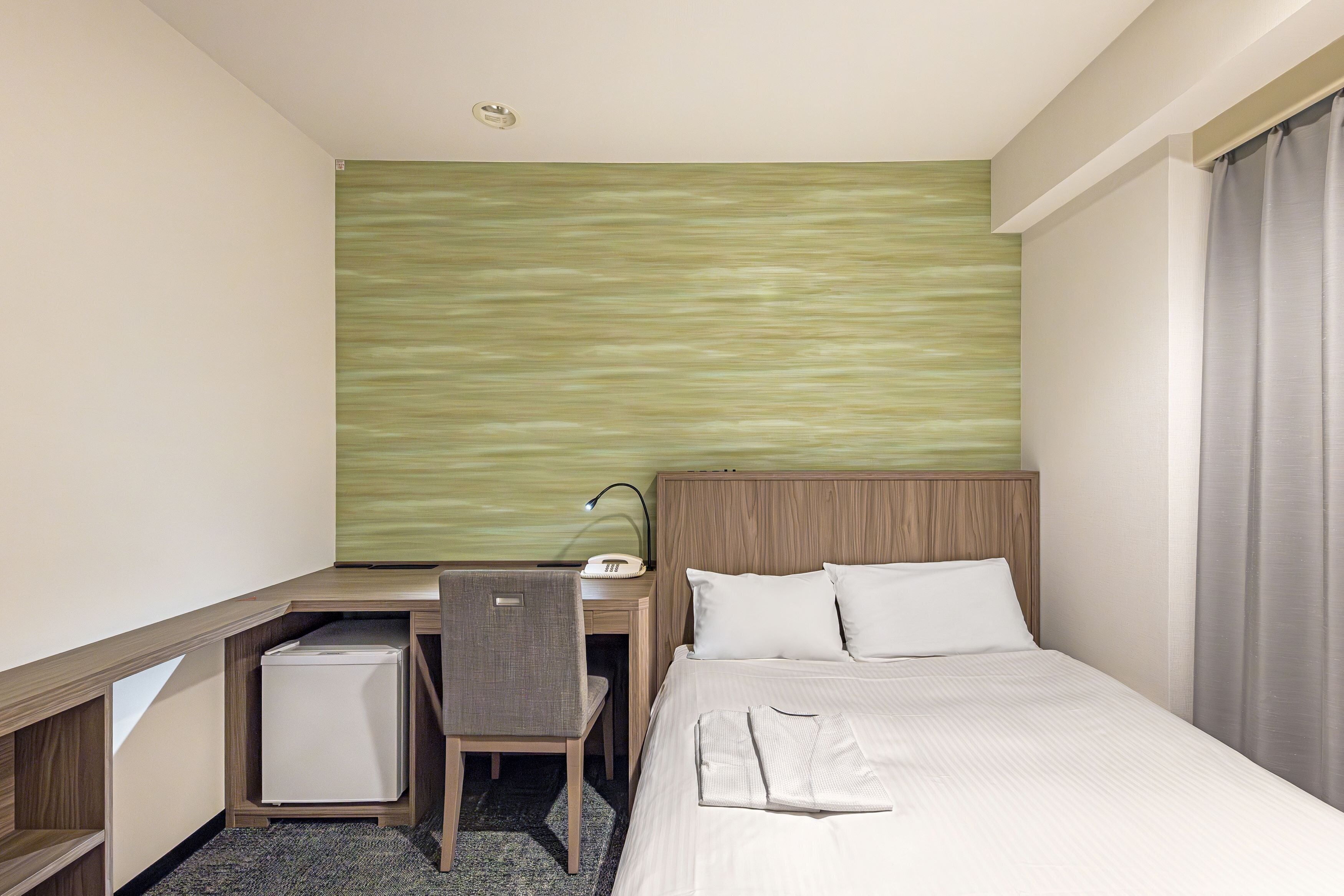 Quarto de hotel com cama, escrivaninha, cadeira e um pequeno frigorífico.