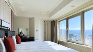 A hotel room with a large bed, a desk, and a view of a cityscape through a large window.