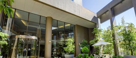 Entrada de hotel con puerta giratoria, fachada de vidrio y zona de estar exterior con plantas en macetas.