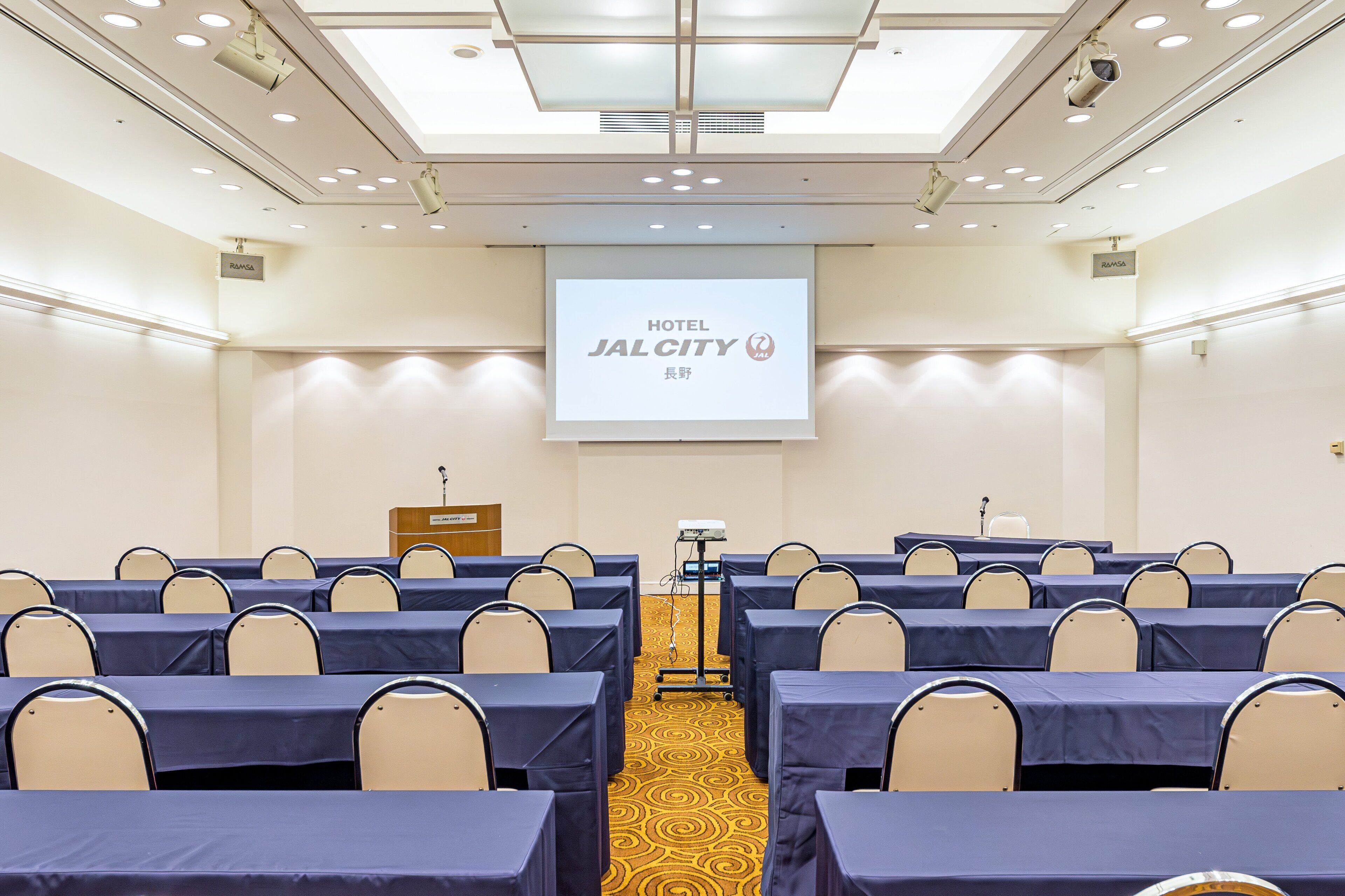 A conference room with rows of tables and chairs, a podium, and a screen displaying 'HOTEL JALCITY'.