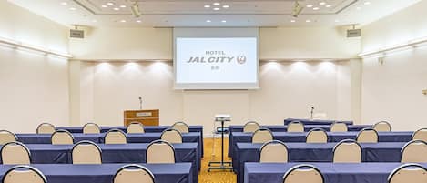 A conference room with rows of tables and chairs, a podium, and a screen displaying 'HOTEL JALCITY'.