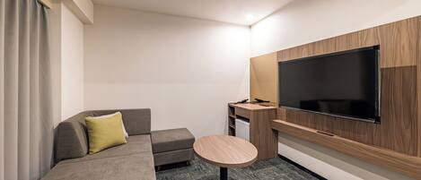 Habitación de hotel moderna con un sofá gris, una pequeña mesa redonda, un televisor montado en la pared y un escritorio de madera.