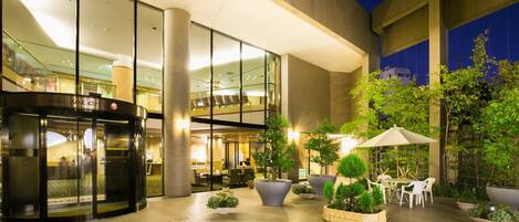 Entrada de hotel con puerta giratoria, zona de estar interior y patio exterior con mesas y sillas.