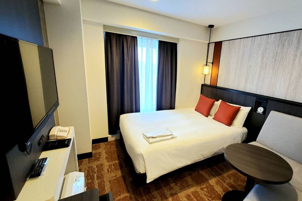 Et hotelværelse med seng, skrivebord med telefon, stol, lille bord og et ur på væggen.