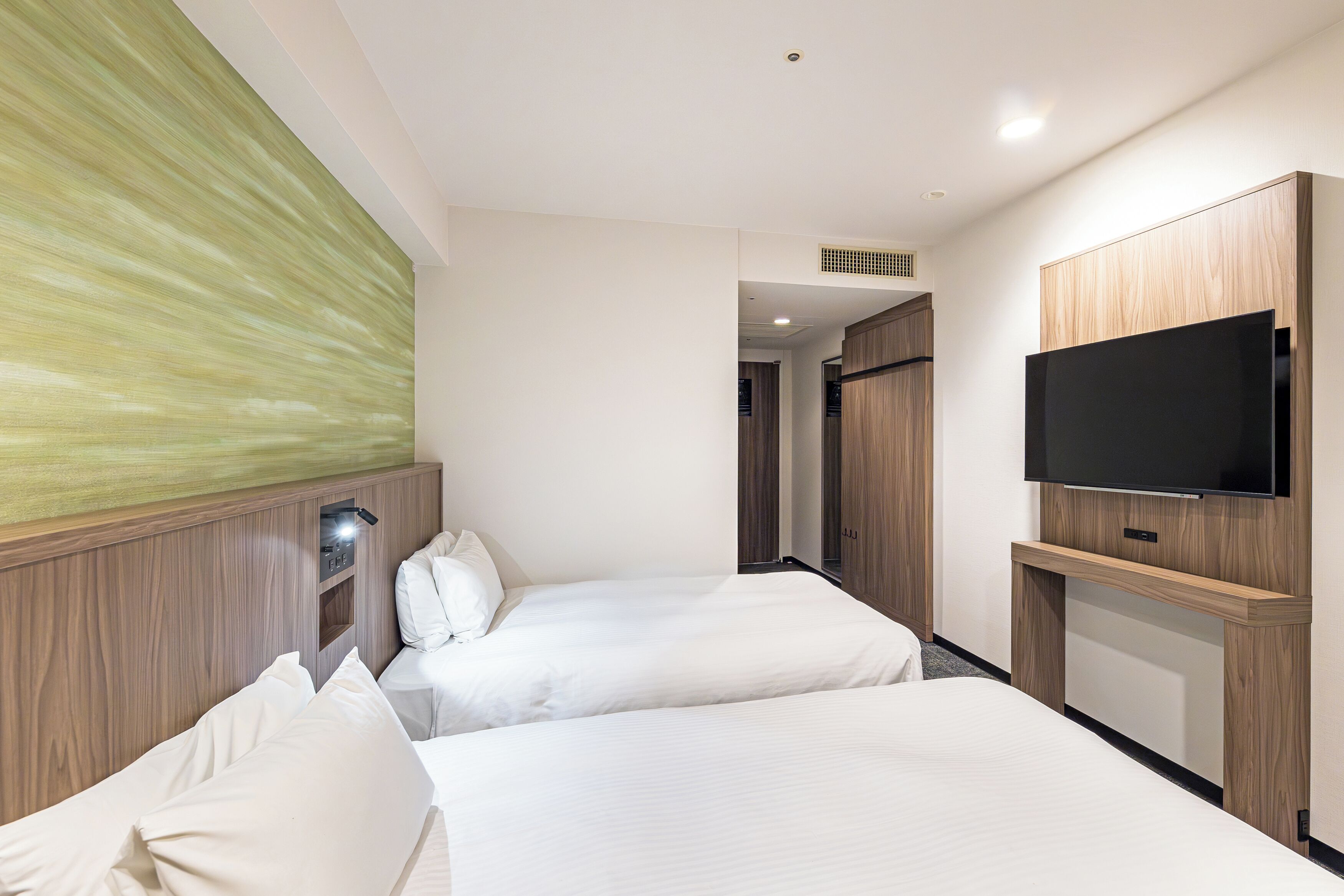 A hotel room with two beds, a flat-screen TV mounted on a wooden panel, and a wall-mounted light fixture.