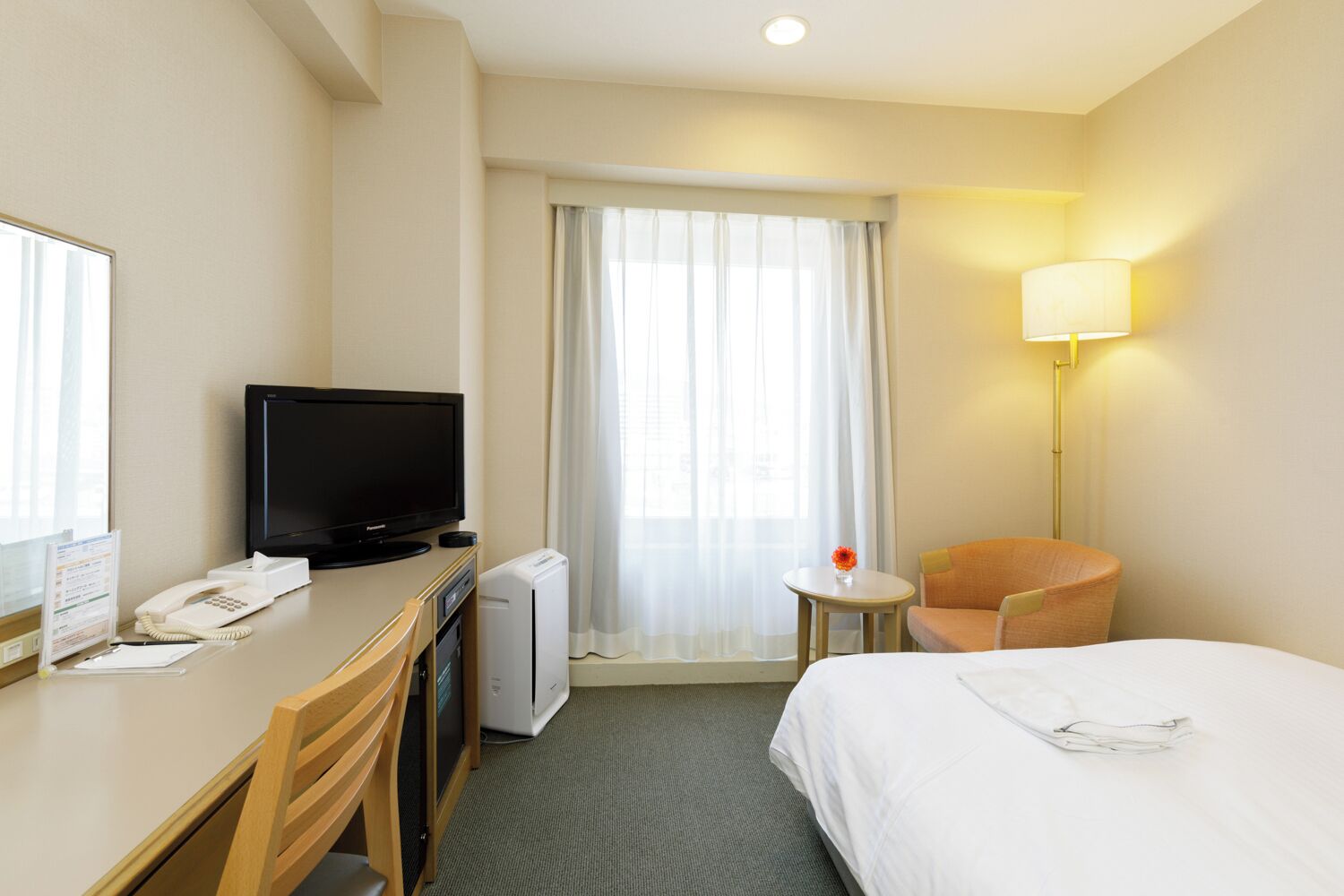 A hotel room with a bed, a desk with a television, a chair, a lamp, and a window with curtains.
