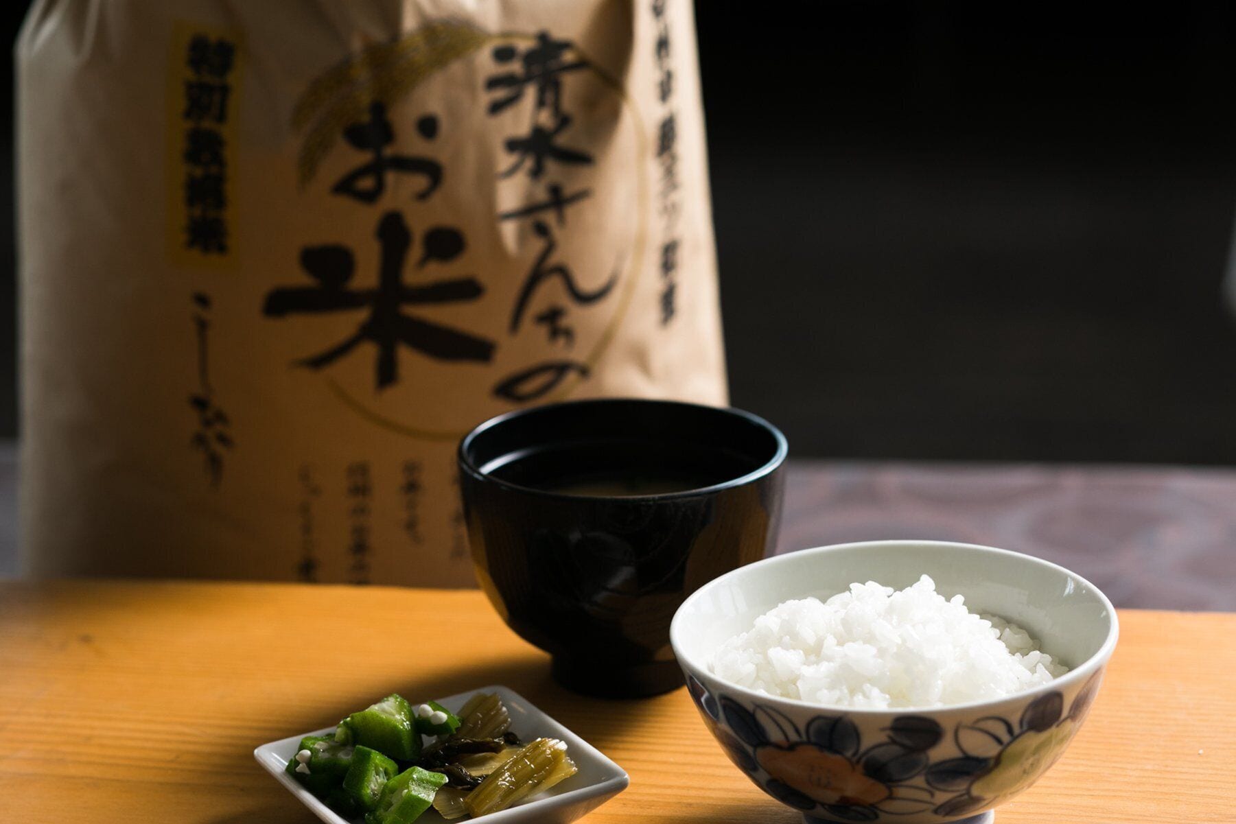 Egy asztalon fehér rizs egy tálban, egy fekete csésze és egy kis tál savanyított zöldségekkel. A háttérben egy papírzacskó látható, japán felirattal.