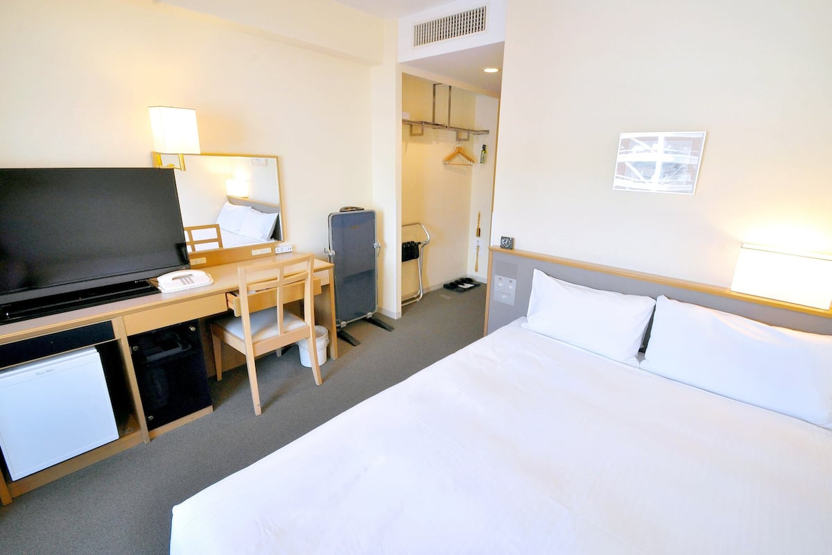 Et hotelværelse med seng, skrivebord med fjernsyn, minikøleskab, stol og knagerække.