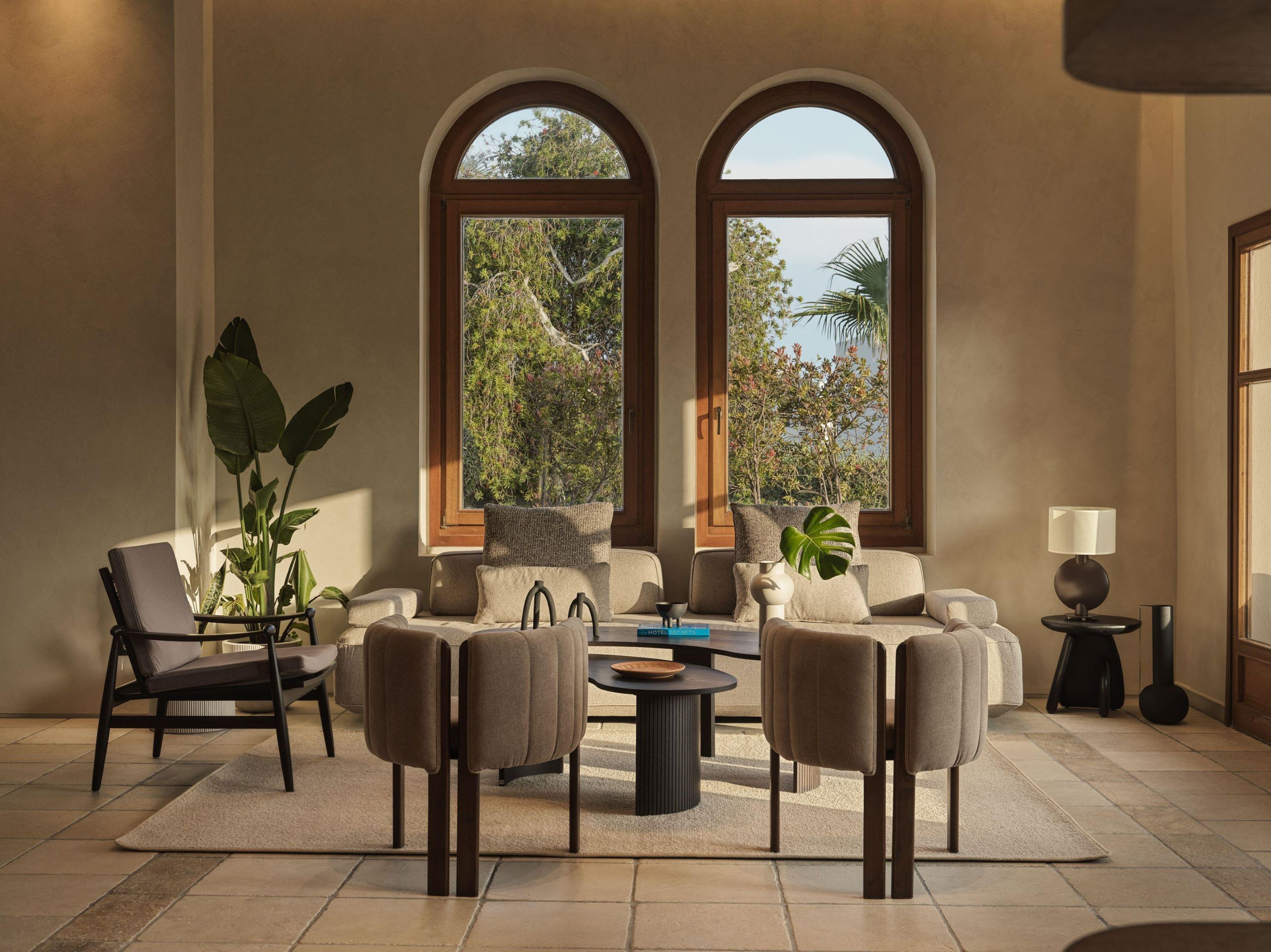 Un salon spacieux comprenant un ensemble de canapés, une table basse et deux fauteuils. De grandes fenêtres à arc, une plante en pot et une lampe posée sur une table d’appoint complètent l’ensemble.