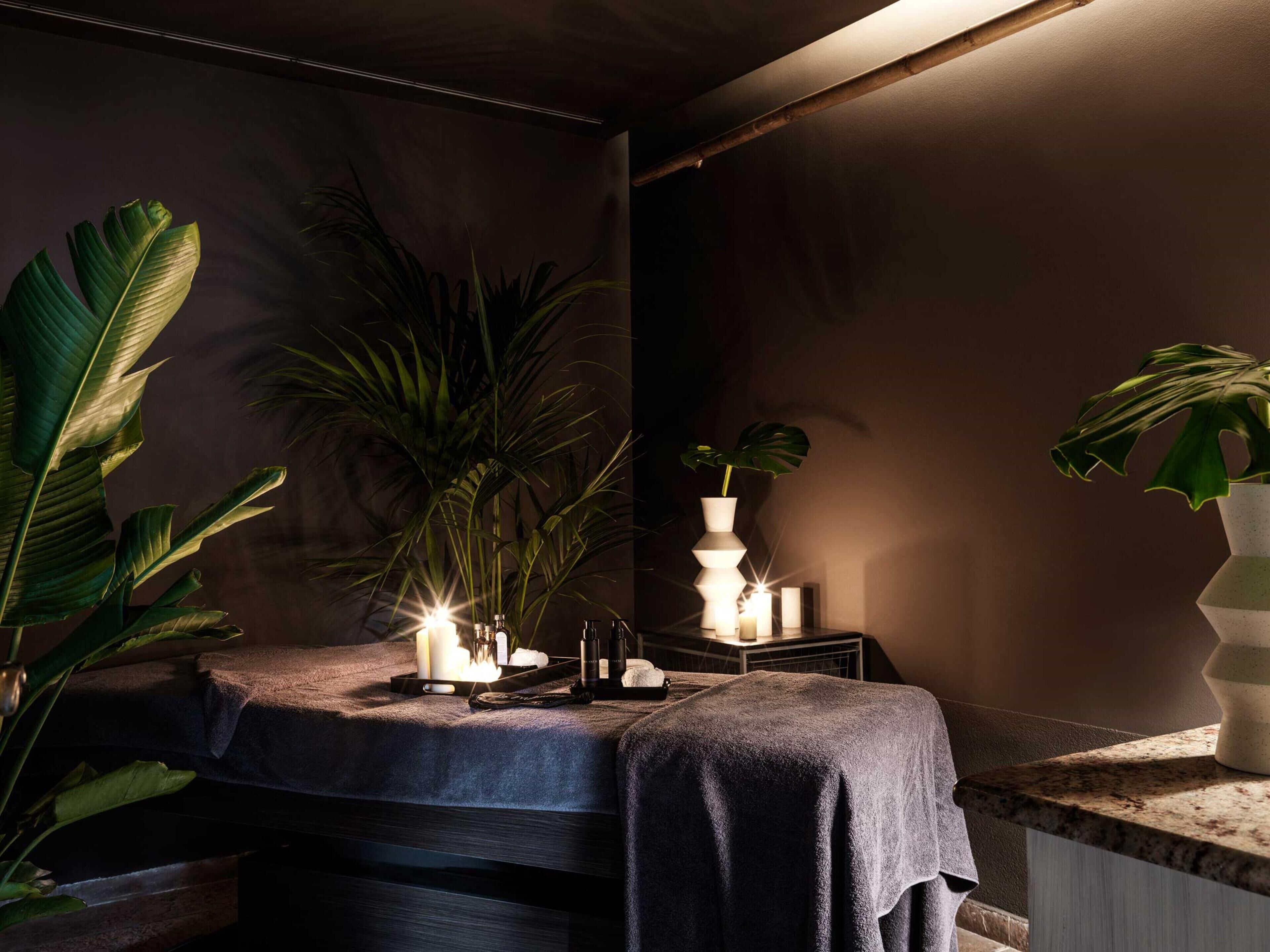 Un área de spa con una camilla de masaje, velas y plantas verdes grandes.