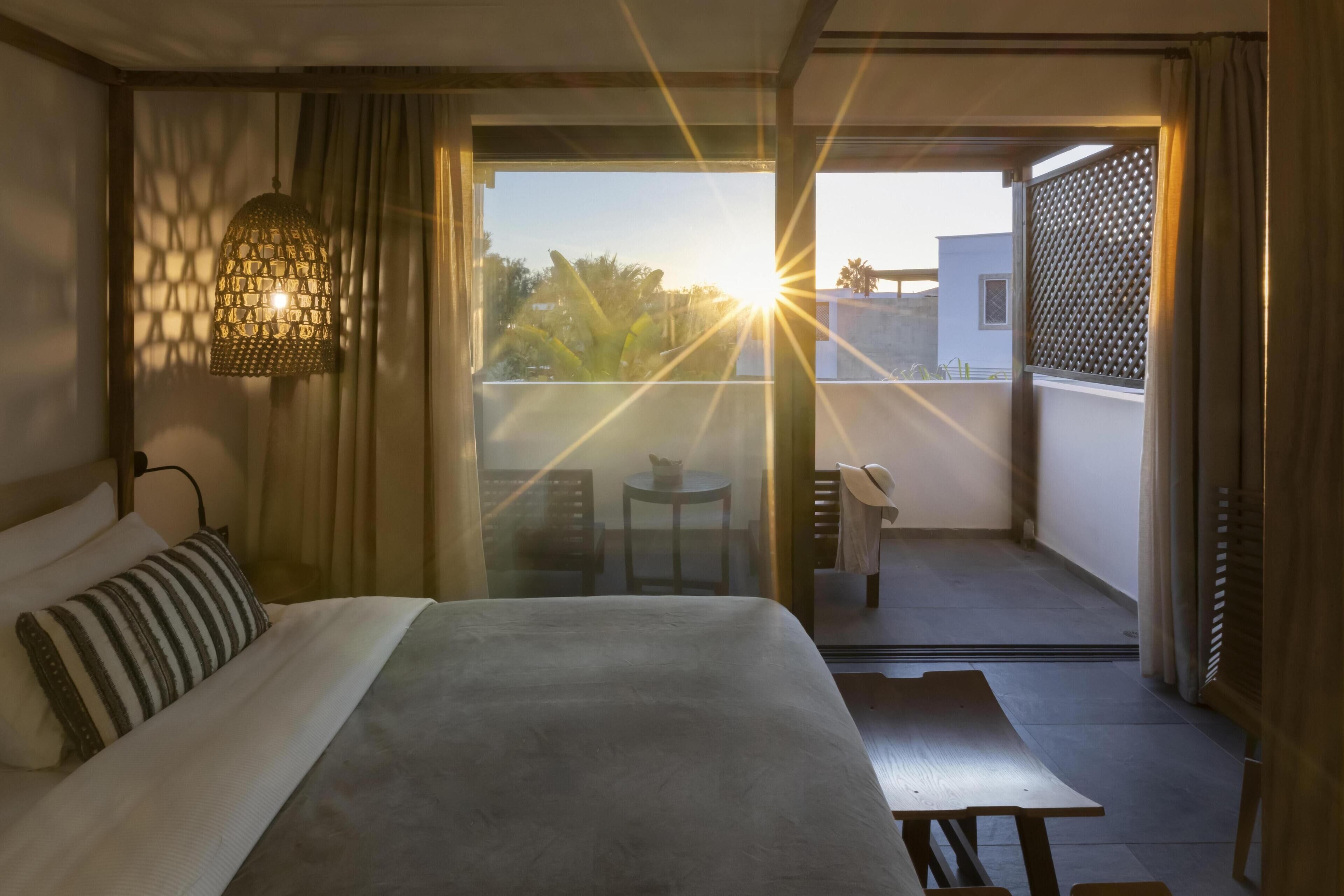 A hotel room with a bed, a hanging light fixture, a balcony with a table and chairs, and a view of the outside.