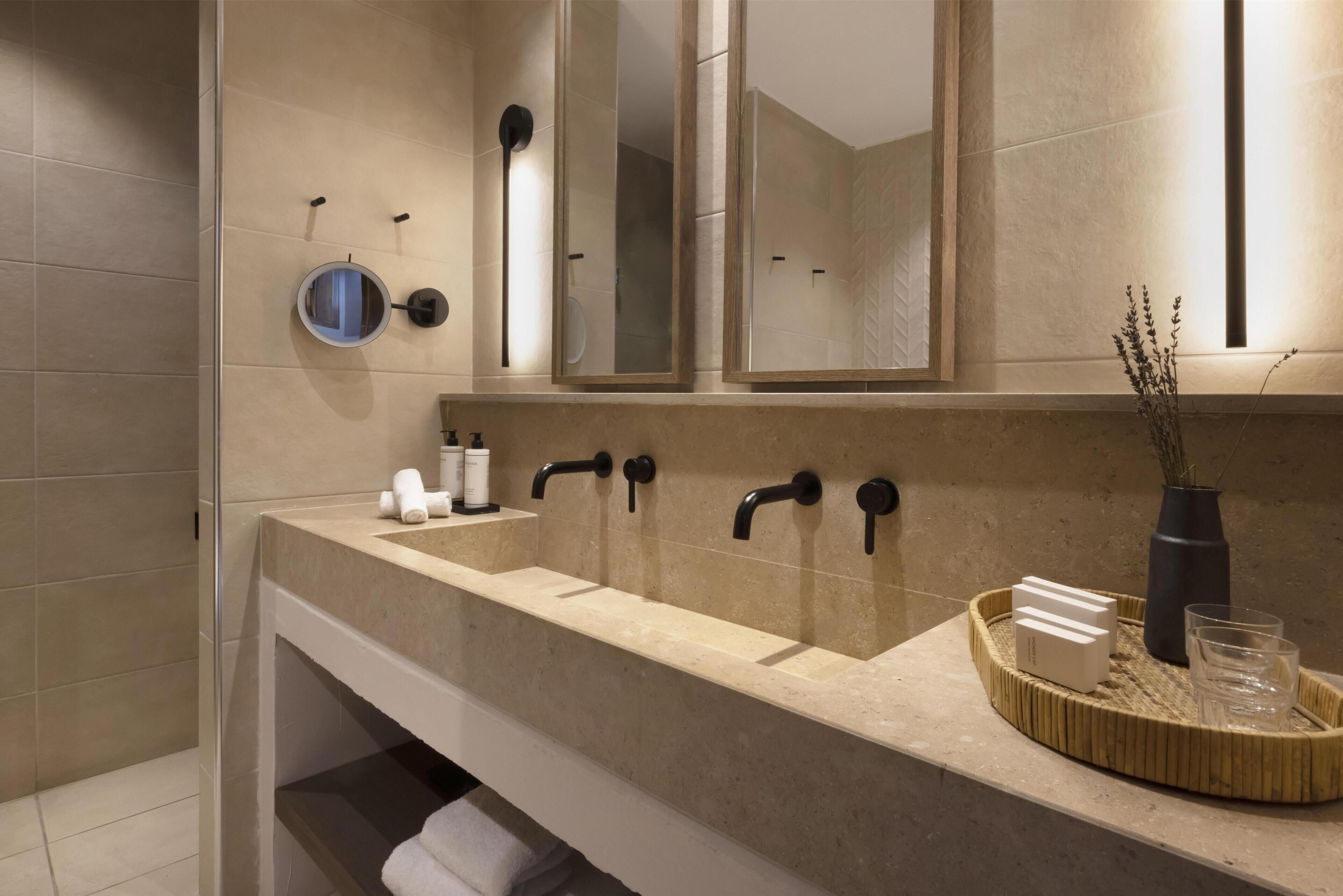 A modern bathroom with a beige tiled wall, a large mirror, a sink with black faucets, and a shelf with toiletries.