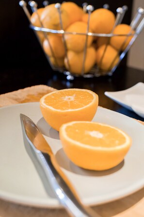 A plate with two halves of an orange and a metal fruit basket in the background. - Parc Hotel (Carvin)
