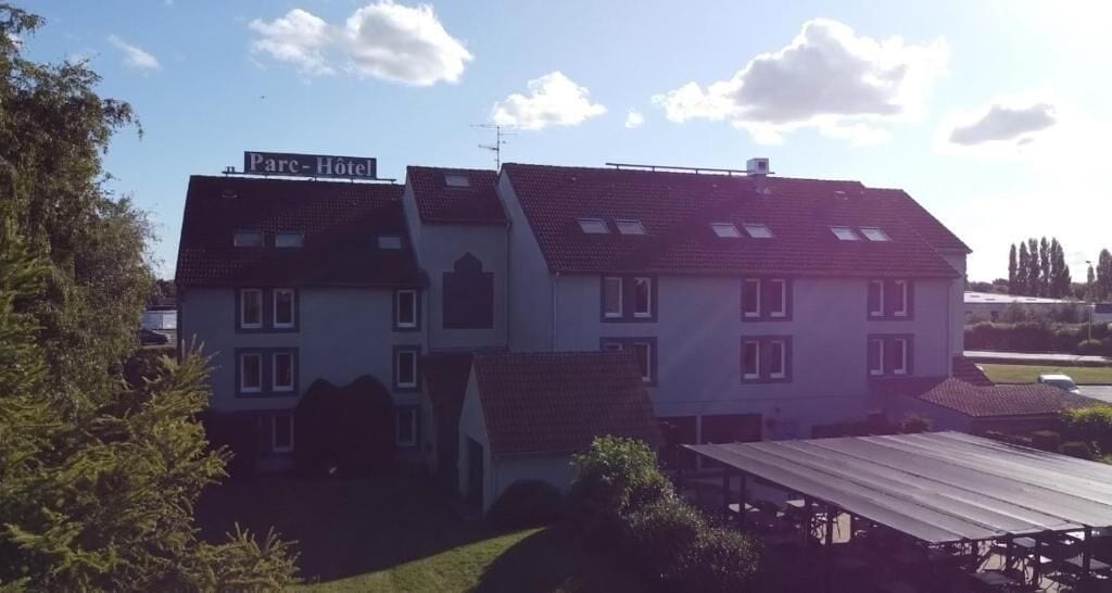Een hotel met een bordje met de tekst 'Parc-Hôtel' op het dak, omringd door groen en een heldere lucht.