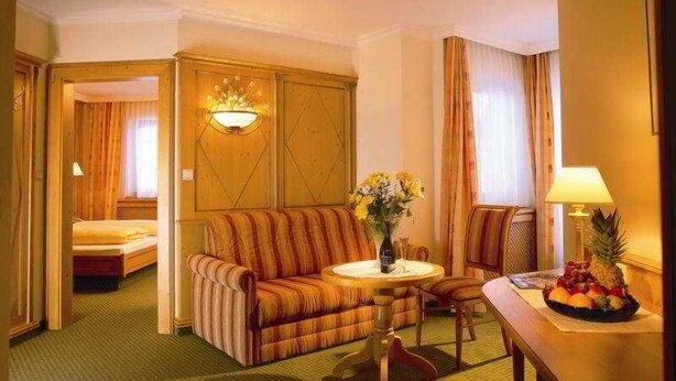 A hotel room with a sofa, a small round table with a vase of flowers, a dining table with fruit, and a bed in the background. - Austria Bellevue (Soelden)