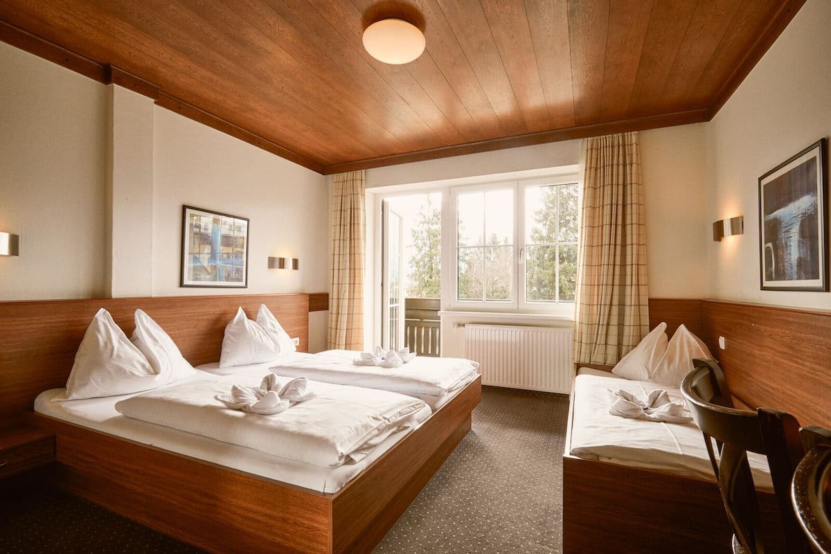 Hotelkamer met twee bedden, houten hoofdborden, een groot raam met gordijnen, een radiator en kunstwerken in lijsten aan de wanden.