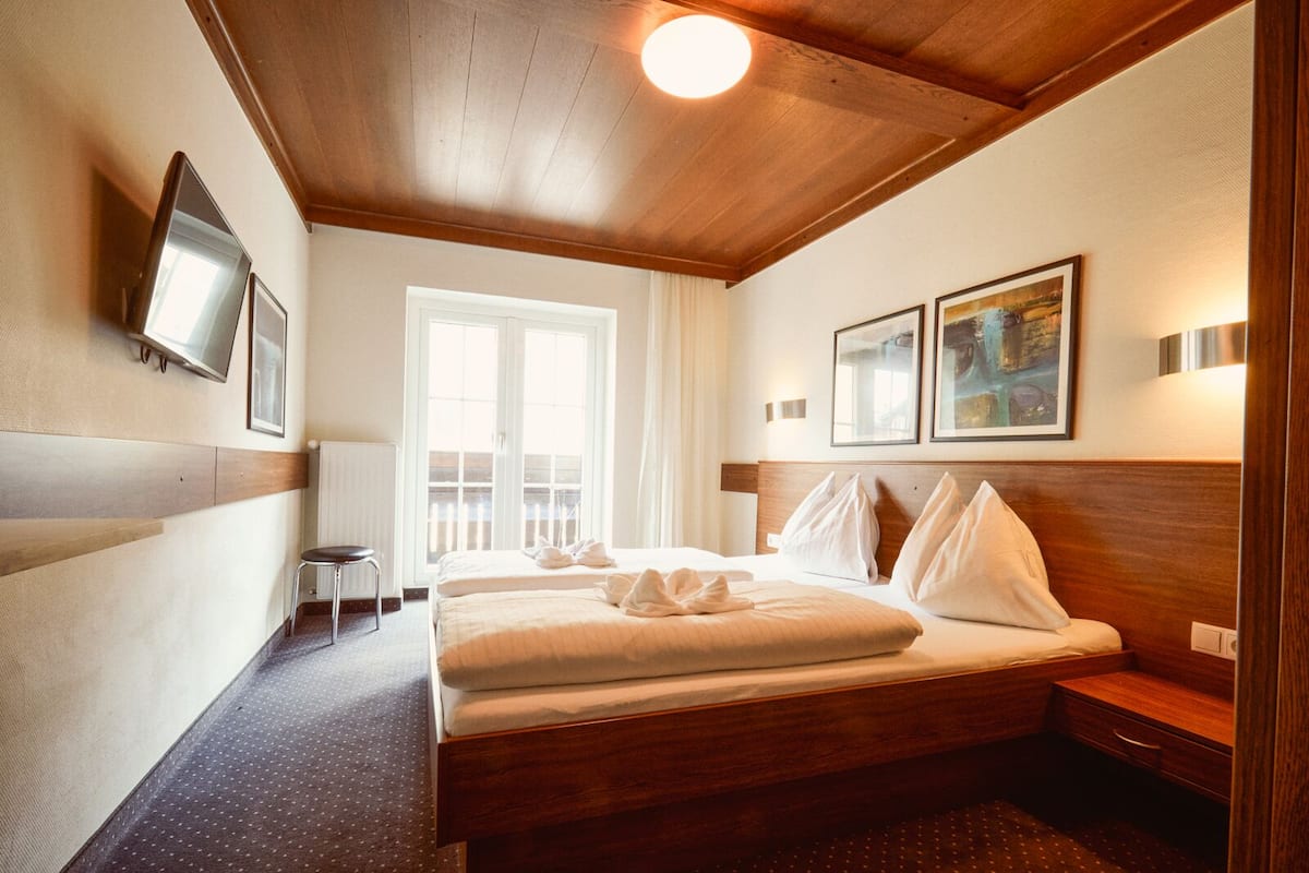 Hotelkamer met een houten plafond, een bed met witte lakens, een kleine tafel en een stoel. Er is een raam met gordijnen, een aan de muur bevestigde televisie en twee ingekaderde foto's boven het bed.