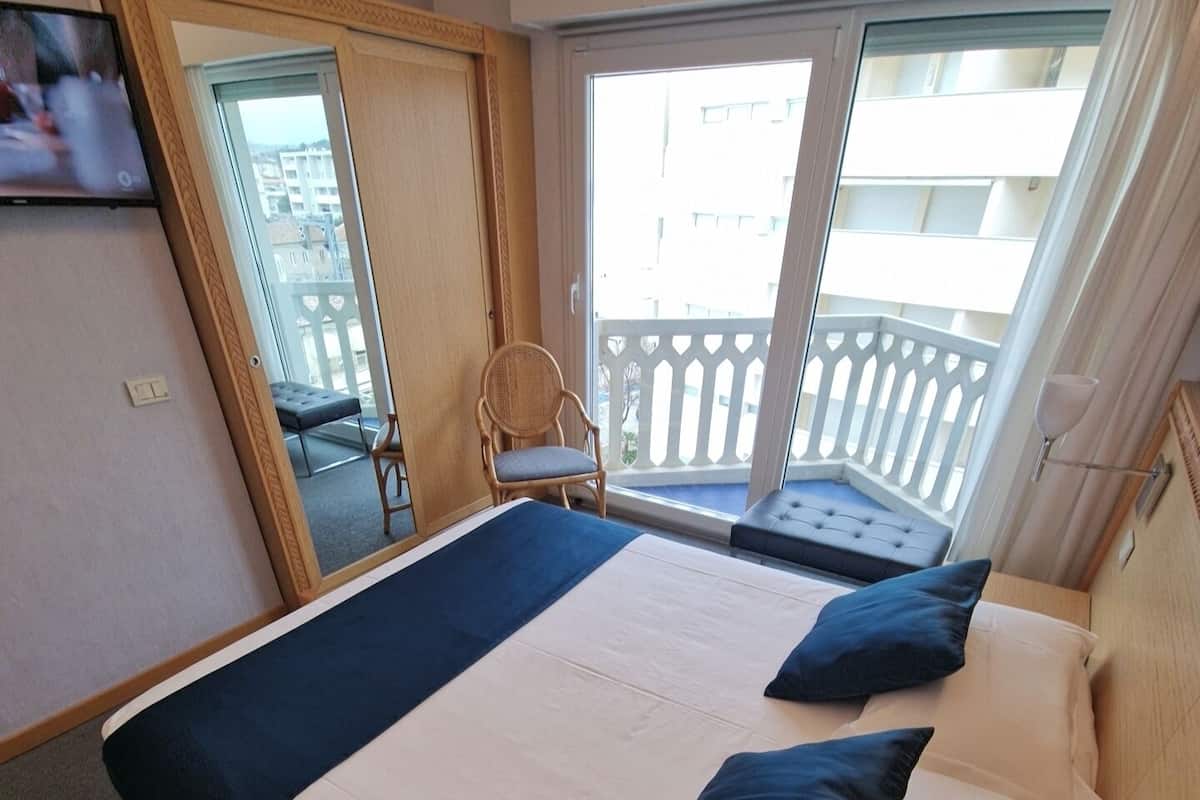 A hotel room with a bed, a chair, a balcony with a railing, and a television on the wall.