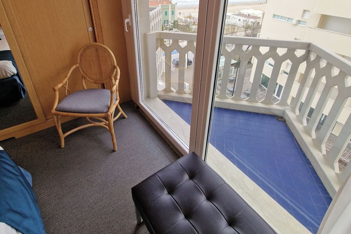 A balcony with a view of the beach and buildings, a wicker chair, and a black cushioned bench.
