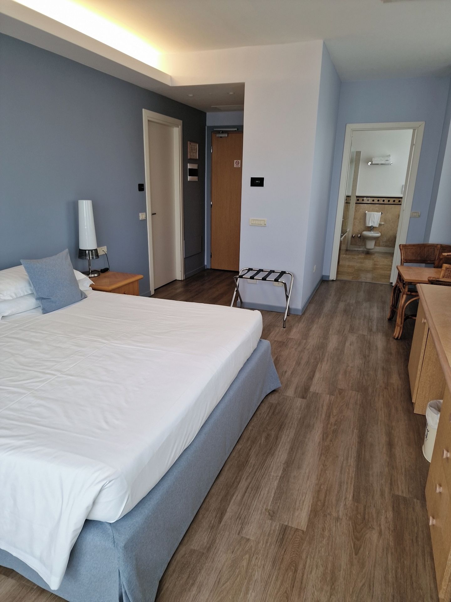 A hotel room with a large bed, a bedside table with a lamp, a wooden floor, and a bathroom visible through an open door.