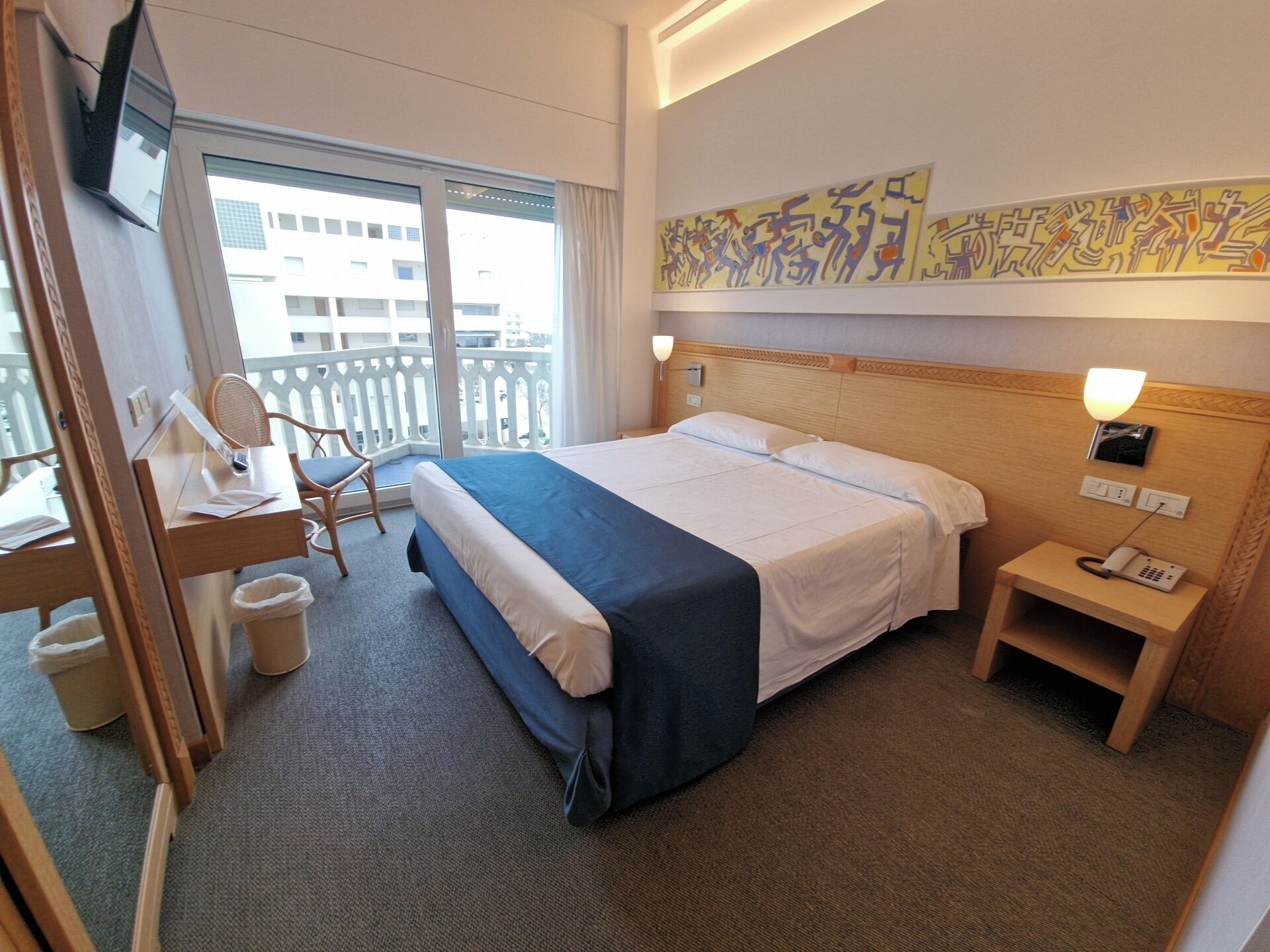 Habitación de hotel con cama, escritorio con silla, mesita de noche con lámpara y ventana con vista a edificios.