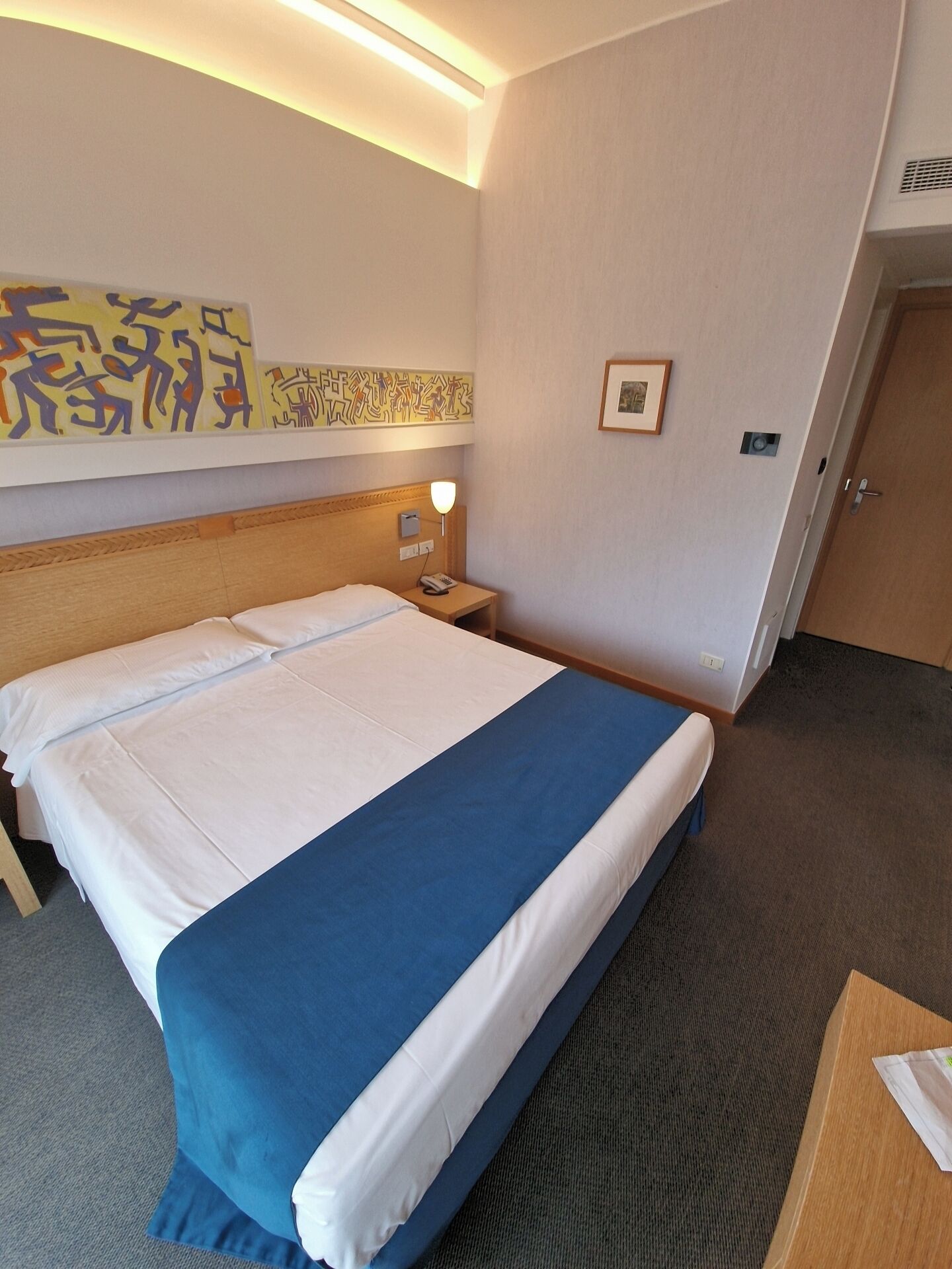 Een hotelkamer met een groot bed, een bedlampje, een klein tafeltje en een muur met abstracte kunst.