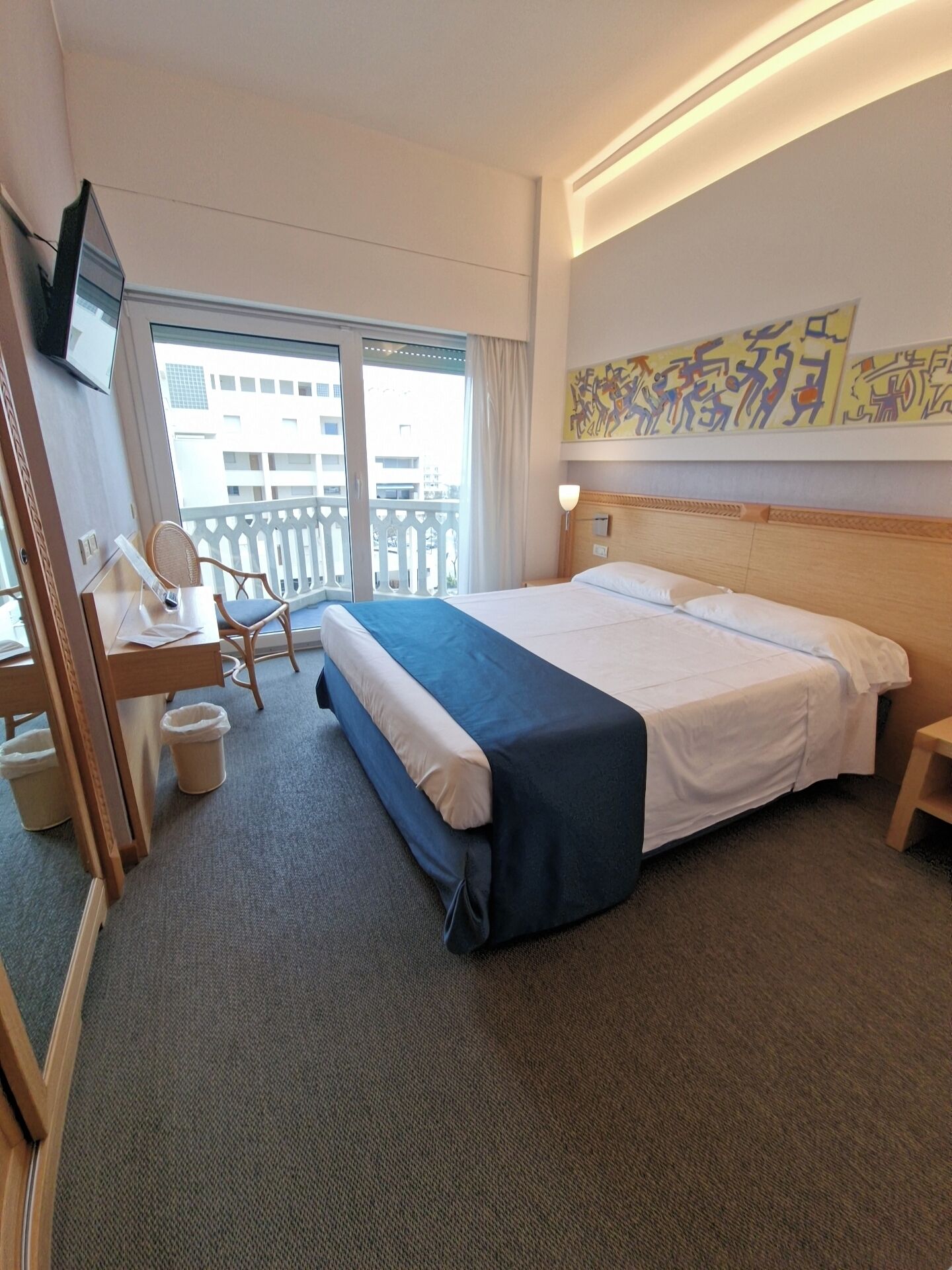 Quarto de hotel com cama, escrivaninha, cadeira, televisão, varanda com vista para edifícios e um quadro na parede.