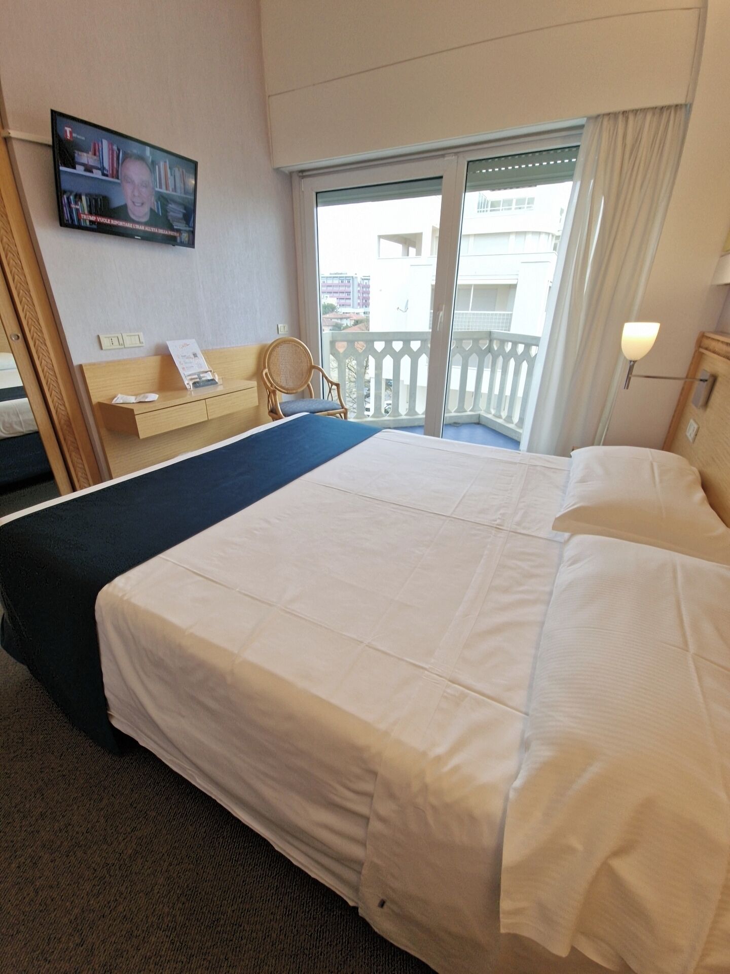 Una camera d'albergo con un letto, una sedia, una scrivania, un televisore e un balcone con ringhiera.